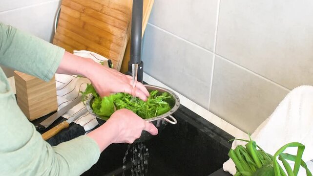 A person is washing various fresh greens in a sink. They hold a colander filled with the greens under running water to clean them. The kitchen is bright and tidy