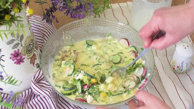 A woman makes okroshka, a cold summer salad. She combines potatoes, radishes, cucumbers, chicken, greens, eggs, and kefir in a bowl. This dish is great for hot days