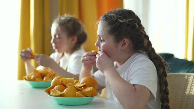 Two girls eating fresh orange slices at table. Child enjoys citrus fruit snack. Girl with braid eats oranges from bowl. Kids have healthy vitamin meal. Children eat fruit together in bright room.