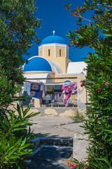 Sunlit Greek church with blue domes and white walls, seen through lush greenery and flowers under clear summer sky on Santorini island. © Andrei Tomas