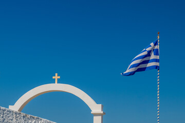 Blue and white Greek flag waving beside a whitewashed arch with cross against clear sky in Santorini, Greece. © Andrei Tomas
