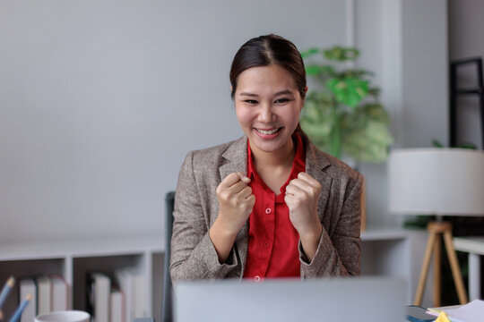Happy young asian business woman celebrating work success or project completion. Clenching fists in excitement while looking at laptop screen in modern office