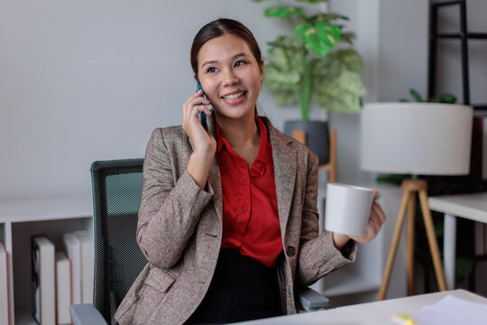 Young businesswoman talking on a smartphone while holding a coffee cup in a modern office, engaged in remote work communication and multitasking