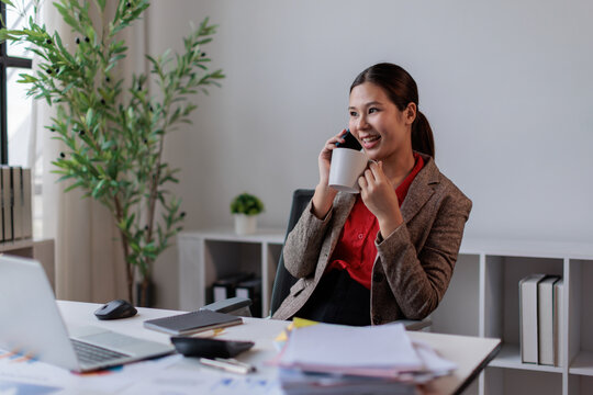 Asian businesswoman communicating by phone call, holding a coffee cup, and smiling while working at her modern office desk with paperwork and technology