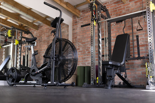 Air bike occupying center in brick-walled gym, showing black fan wheel with power rack, bench, mats