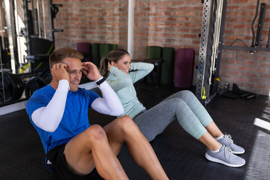 Couple performing abdominal crunches on rubber flooring near squat rack and mirror in athletic wear