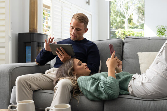 Diverse couple relaxing on grey sofa in living room, man tapping tablet, woman scrolling smartphone