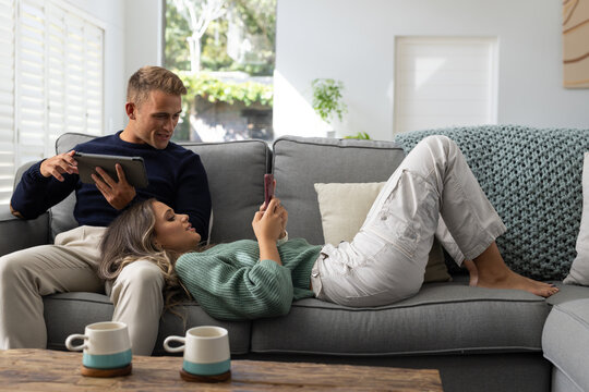 Diverse couple reclining on grey sofa with coffee table, woman holding smartphone, man using tablet