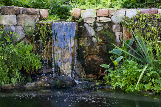 Stacked stone wall is channeling thin waterfall into pond, with moss, ferns, rippling surface
