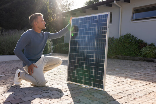 Adult man kneeling and inspecting photovoltaic panel on stone-paved driveway by house exterior