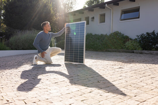 Adult male kneeling on sunlit brick driveway holding and inspecting photovoltaic solar panel