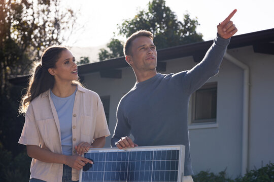 Diverse couple standing outside single-story house holding solar panel, pointing toward roof
