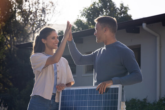 Diverse couple holding solar panel and high-fiving while standing by house exterior