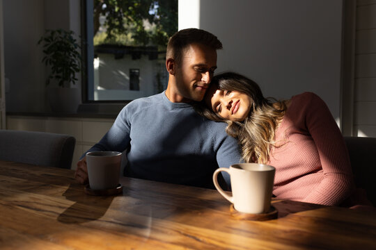 Diverse couple sitting close on bench at wooden dining table near large window with ceramic mugs