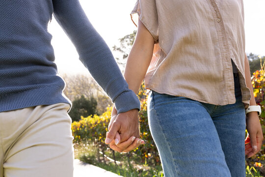 Couple walking holding hands wearing casual clothes on sunlit path with shrubs, showing white watch