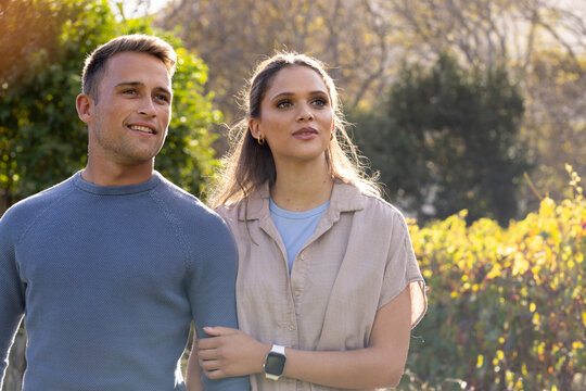 Diverse couple standing arms linked in sunlit garden showing smartwatch and fitted sweater