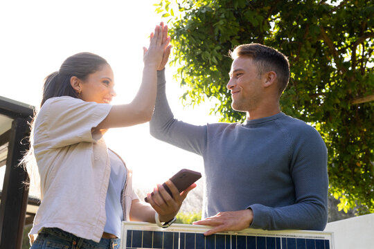 Diverse couple high-fiving, man holding solar panel while woman holding smartphone in backyard