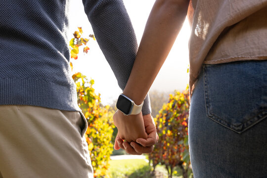 Couple holding hands while walking in sunny garden path, showing smartwatch and textured clothing