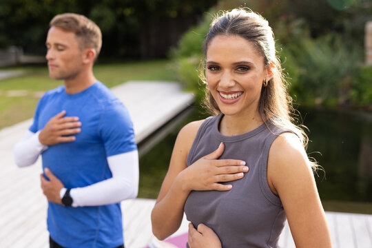 Diverse partners in sportswear and smartwatch doing breathing on wooden dock by pond with pink mat