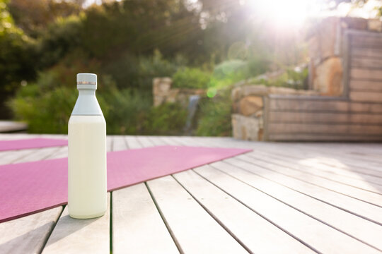 Pale reusable water bottle is sitting on light wooden deck near pink yoga mat, small waterfall