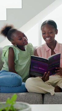Vertical video: Opening purple book teen sister reading to sister on gray sofa by window for fun