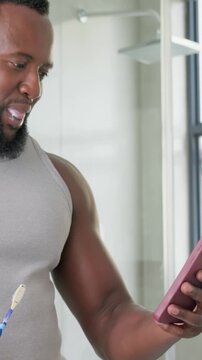 Vertical video: Brushing man in grey tank reading texts at home holding toothbrush and pink phone