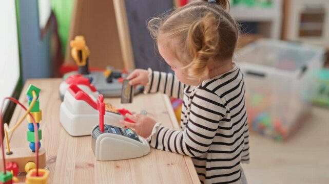 Little blonde girl presses buttons on a toy register and holds a play card while standing at a wooden table in a building; curiosity learning play.