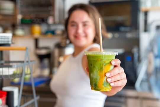 A woman barista holds out a green iced mango matcha latte with plant-based milk in a clear plastic cup with a straw inside a coffee shop.