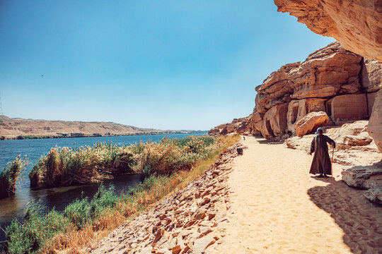 Edfu, Egypt - 02 March 2020: View of the Nile riverbank with rock-cut shrines, a sandy path, and a man in traditional dress standing near the water.