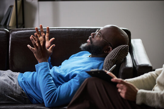 Adult Black man reclining and speaking during therapy session while female therapist taking notes, conveying counseling and mental health support. For relationship care marketing