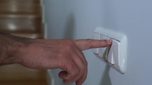 A close-up video of a man's hand pushing a white light switch on the wall to turn on the lights in a modern house hallway.