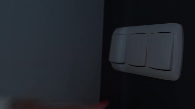 Close-up view of a female hand pushing a white light switch button to turn off the lights, leaving the room in darkness to save power.