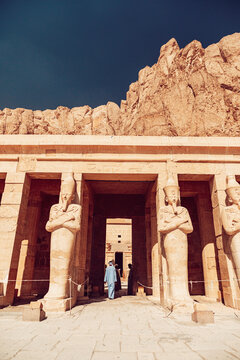Luxor, Egypt - 04 March 2020: View of the Mortuary Temple of Hatshepsut with Osiride statues and people walking through the entrance against rugged limestone cliffs.