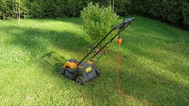Eco-friendly electric lawn mower with orange cable on green grass. Eco lawn care tool protecting nature. Electric lawn mower with orange cable on green grass in backyard. Lawn care and gardening tool.
