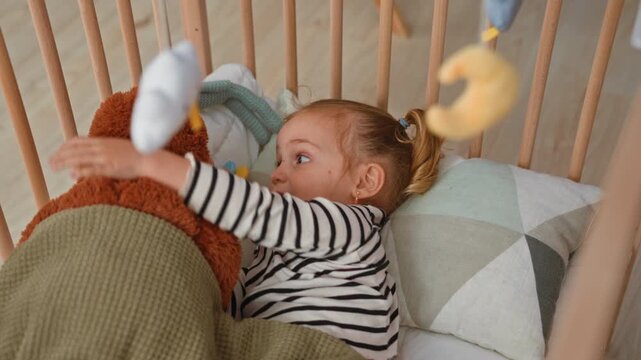 Blonde little girl in wooden crib grips rail and reaches for hanging plush mobile toys in building; curiosity play.