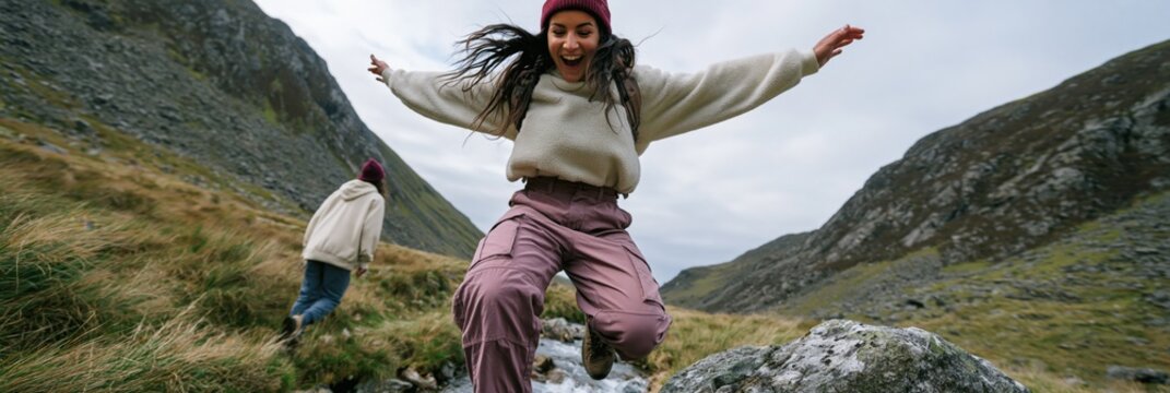 Young woman jumping over a rocky brook in a mountain valley while a companion walks behind for adventure banner background and outdoor travel poster