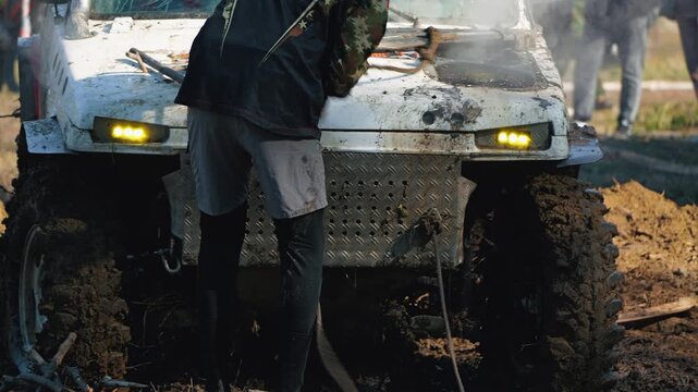 A close-up of a powerful 4x4 SUV, covered in mud and stuck in thick slush. A co-pilot lays a winch cable on the hood of the smoking car during an intense off-road rally. Extreme motorsports