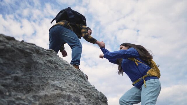 Two hikers with backpacks assist each other while climbing rocky mountain terrain. Adventure partners demonstrate teamwork and support during challenging outdoor expedition under cloudy sky.