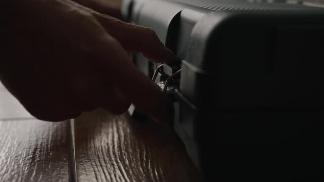 Close-up of male hands unlocking and opening dark plastic toolbox, revealing professional equipment inside and preparing for work on wooden floor in dark interior of room with natural light.