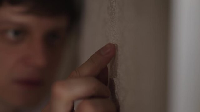 Focused individual applying small pieces of clear adhesive tape onto textured wall, pressing each section with finger in dark interior of room with shallow depth of field. Shooting in slow motion.