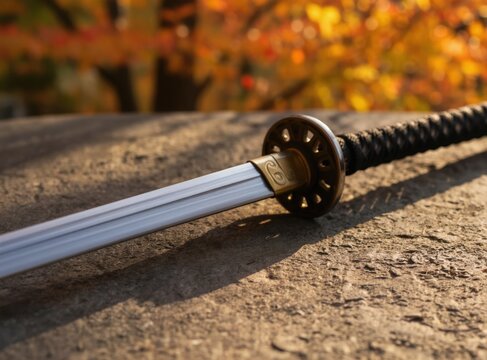 Katana sword rests on stone surface. Blurred orange leaves and sunlight create an atmospheric background. Weapon features sharp steel blade and detailed hilt.