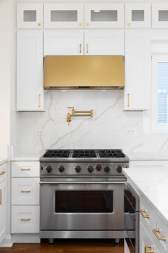 A white kitchen detail with a marble backsplash and countertop, stainless steel oven, and gold range hood, hardware, and pot filler faucet.