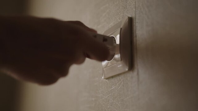 Close-up hand of unrecognizable person inserts white electrical plug into wall socket. Connecting appliance to power source. Concept of electricity, energy consumption, domestic life, slow motion.