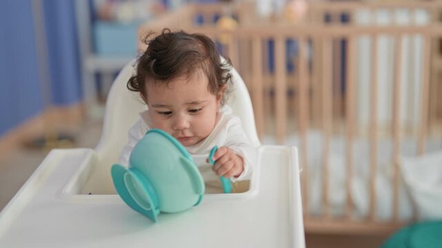 Hispanic girl grabs a teal suction bowl and spoon with her hand while seated in a white highchair in a nursery room inside a building; curiosity.