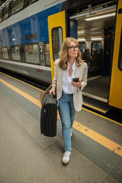 Mature businesswoman waiting for train with suitcase and smartphone at railway station platform