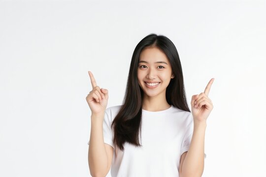 Smiling young Asian woman in white shirt points fingers up. She gestures to present information or indicate a direction against a plain background. Copy space available for text.