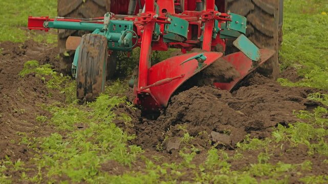 Closeup view of heavy agricultural machinery plowing field, red metal plow turning over dark earth, green grass surrounding furrow, farming equipment in action, rural landscape scene