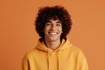 Obraz premium Young man with curly hair smiles. He wears a yellow hoodie against an orange background. His expression is happy and confident. This photo represents joy and youth.