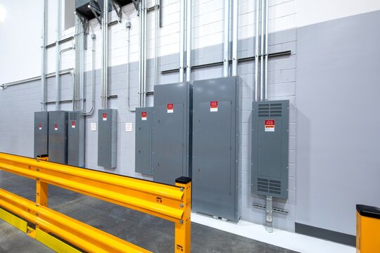 Industrial electrical panels and metal conduits mounted on a gray concrete wall behind a safety guardrail in a warehouse