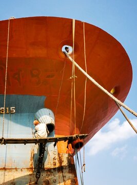 Dhaka, Bangladesh - 03 June 2026: View of workers performing maintenance on the massive orange and blue hull of a ship with ropes and wooden scaffolding under a clear sky.